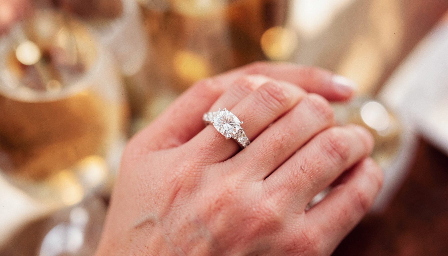 A close-up of a diamond engagement ring on a hand against a soft blurred background, warm champagne and rose-gold tones.