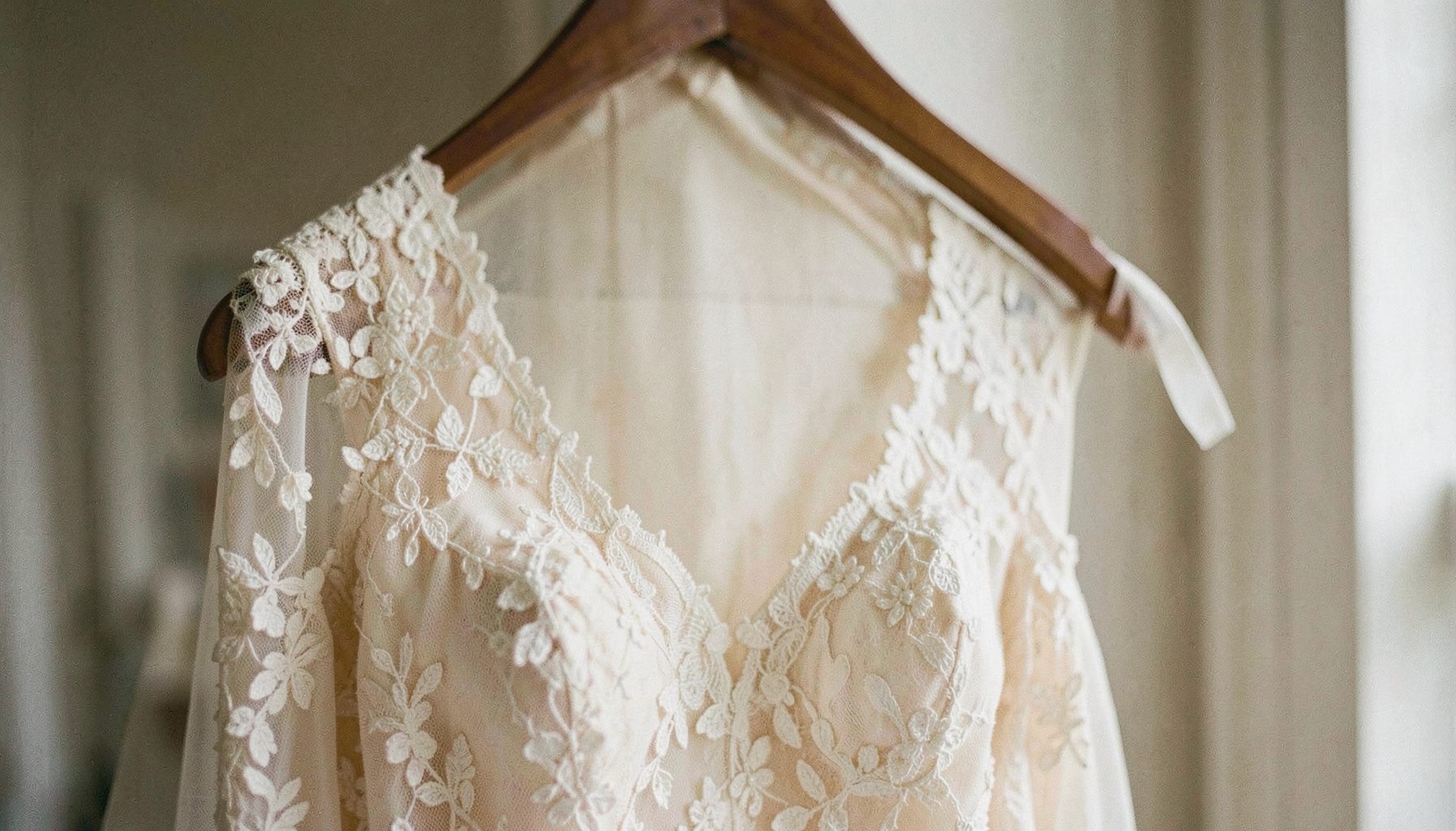 A close-up of a delicate ivory lace wedding gown draped over a wooden hanger, soft natural daylight, cream and blush tones, candid documentary style.